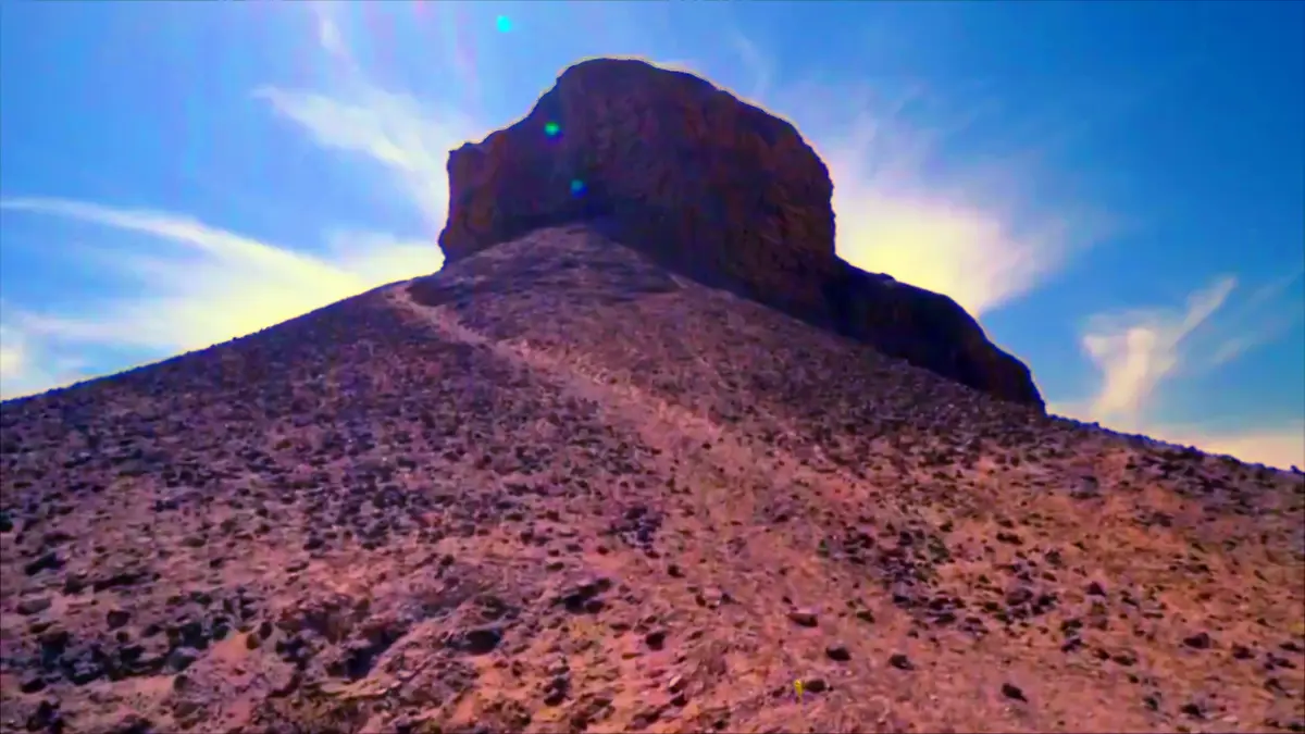 A low-angle shot shows a lone, tiny human figure at the base of a vast, steep, rocky mountain under a bright blue sky with…