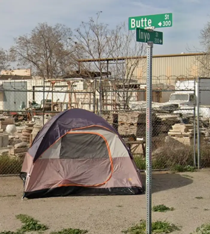 A purple and gray camping tent with orange trim sits on a gravel lot near a chain-link fence, with two green street signs…