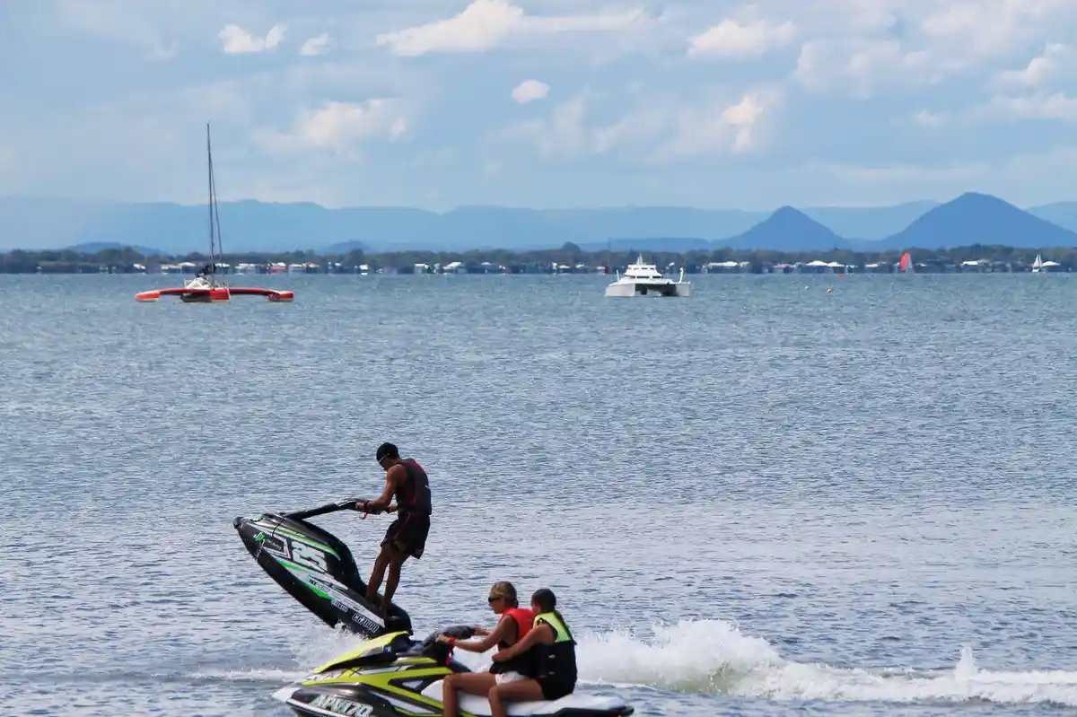 A man stands on the back of a jet ski, lifting it high out of the water while two people sit on another jet ski in front of…