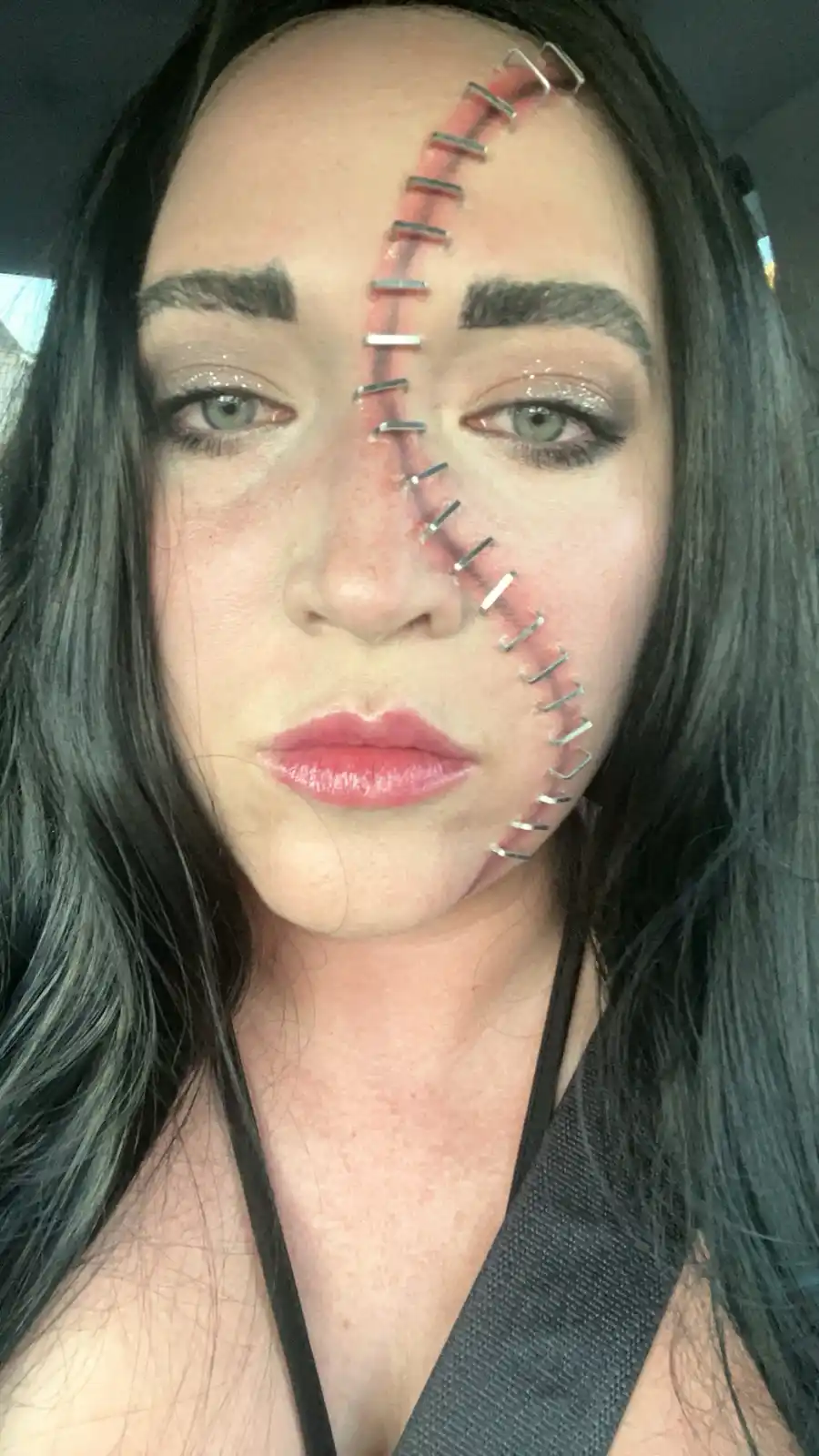 A person with long dark hair and makeup is shown in a selfie, with a prominent, theatrical stitched-up scar running…