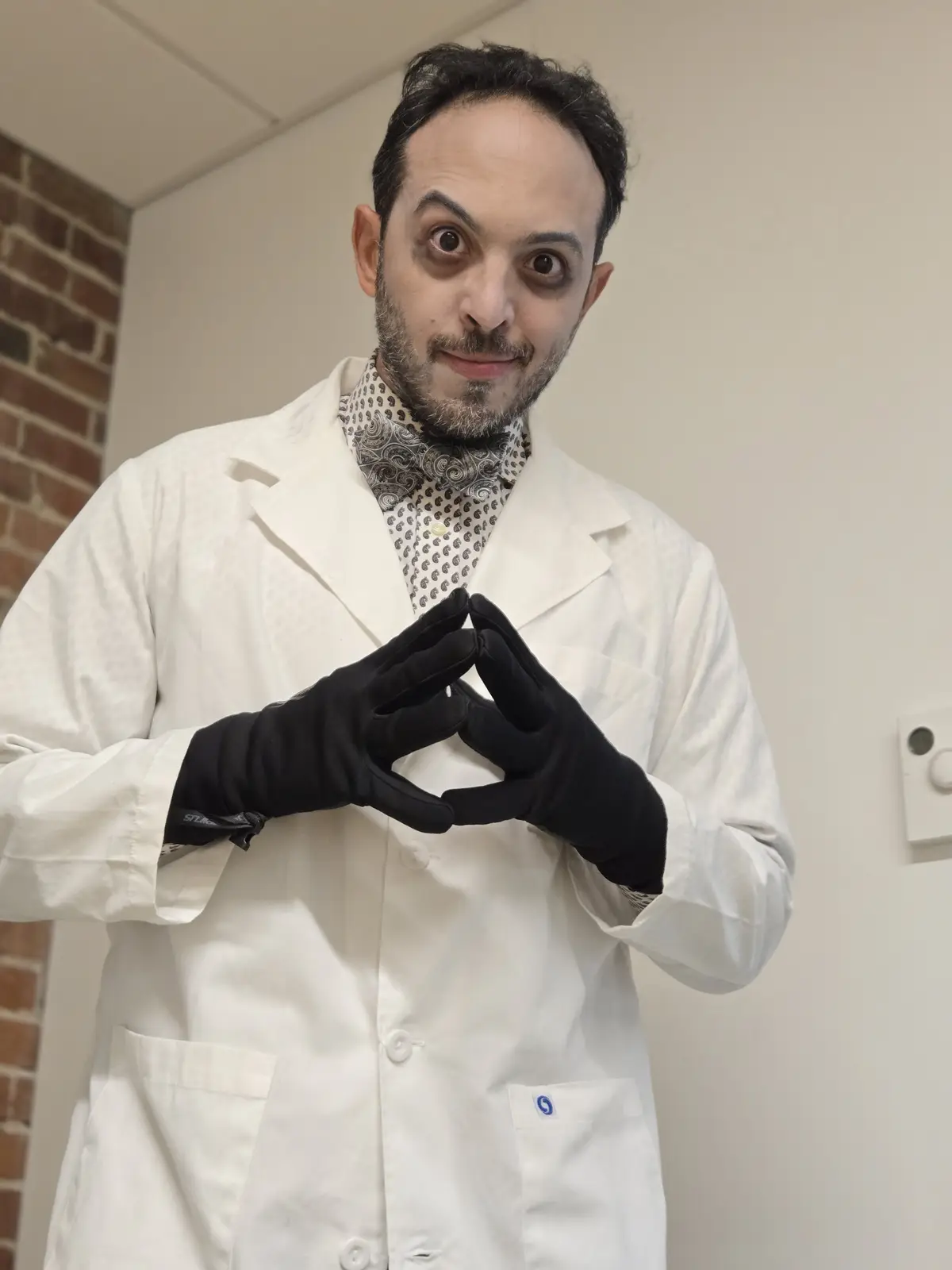A man in a white lab coat and black gloves makes a heart shape with his hands while wearing dramatic makeup, including dark…
