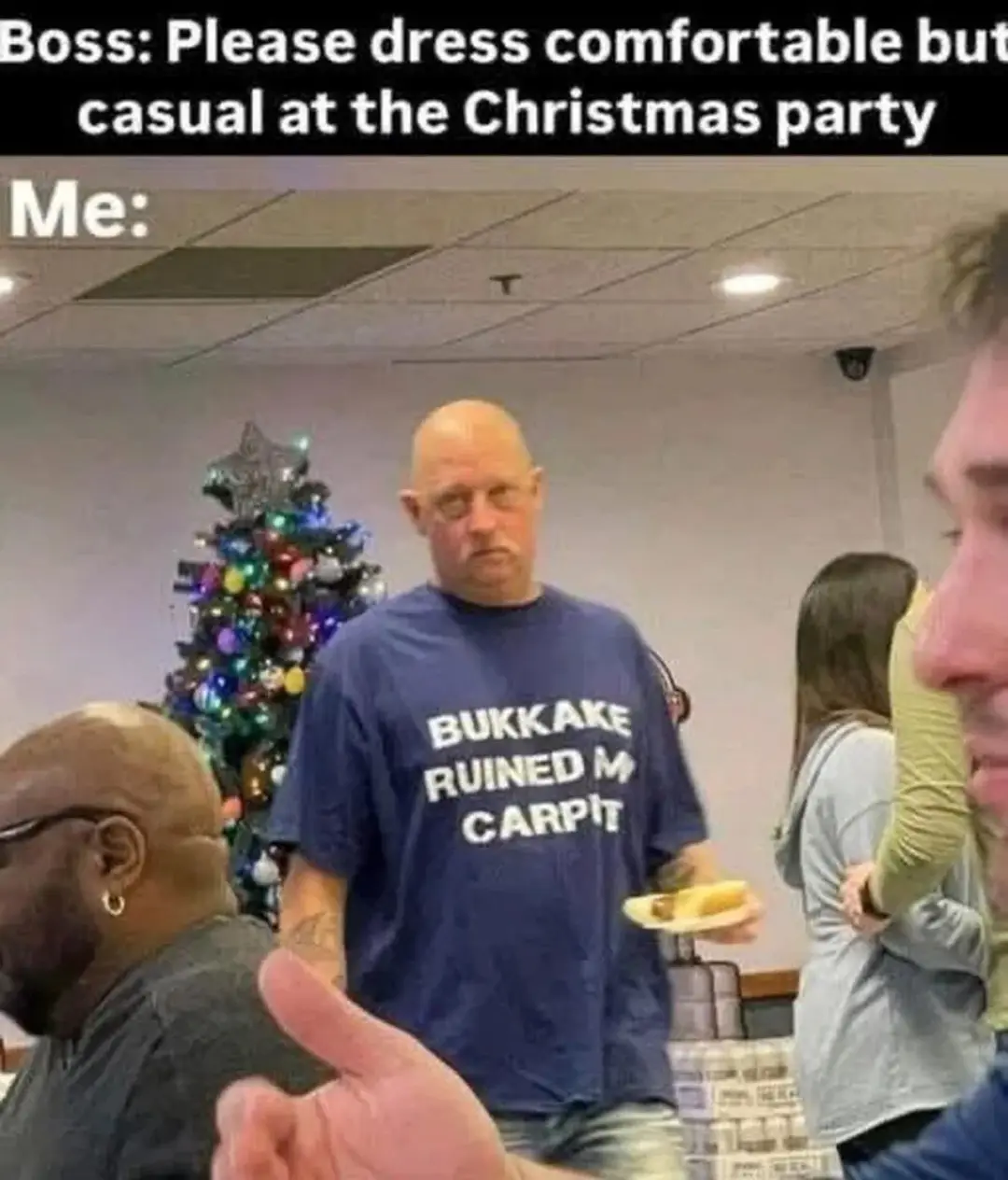 A man in a blue t-shirt with the text 'BUKKAKE RUINED MY CARPIT' stands in a room with a decorated Christmas tree behind…