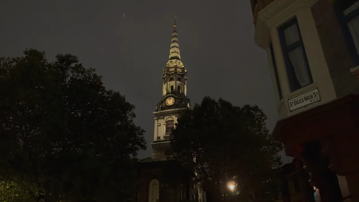 A nighttime photo of a lit church steeple with a clock, partially obscured by trees, beside a building with a street sign…