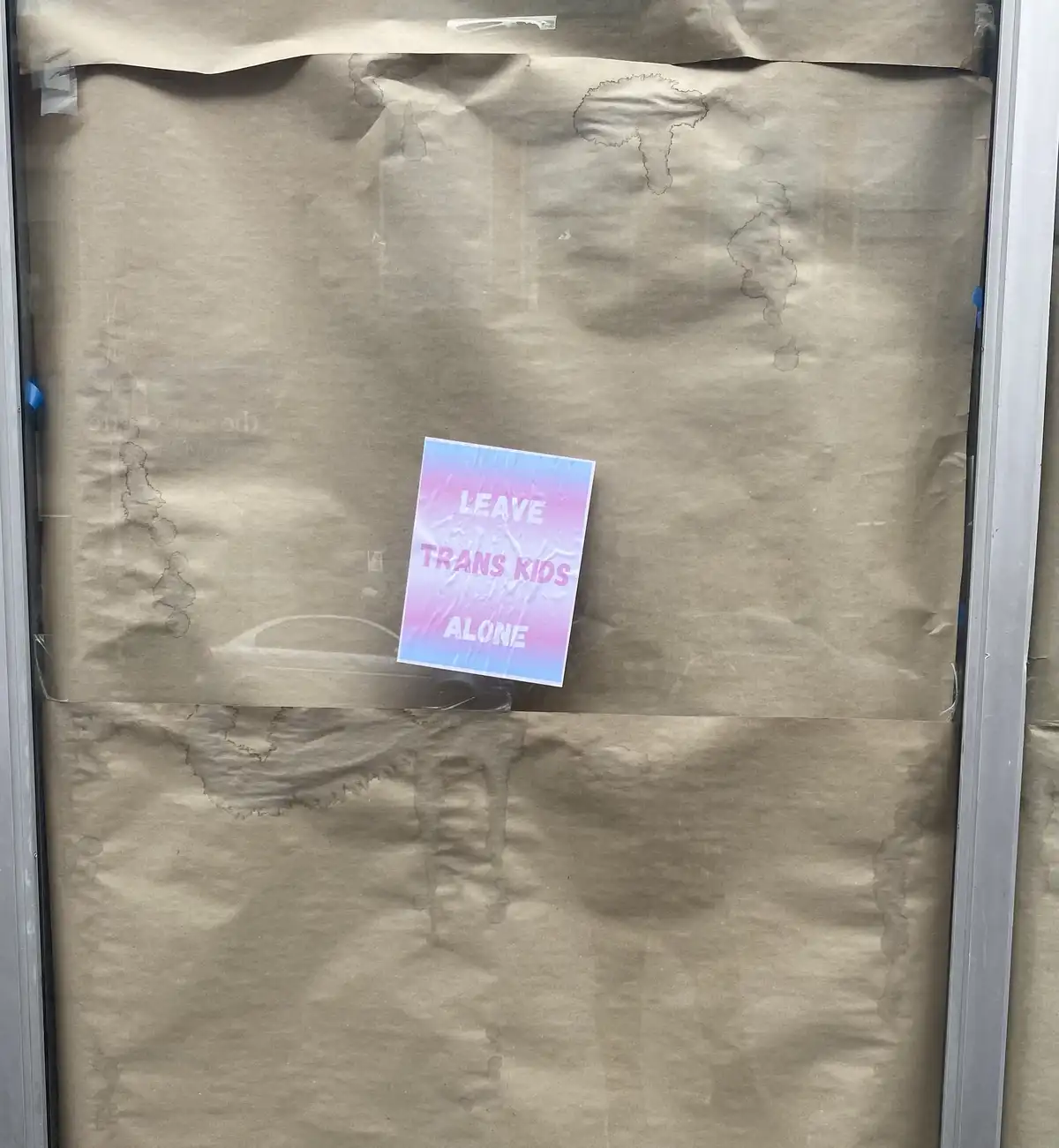 A small, colorful sign with a pink and blue gradient background is taped to a glass door covered with brown paper. The sign…