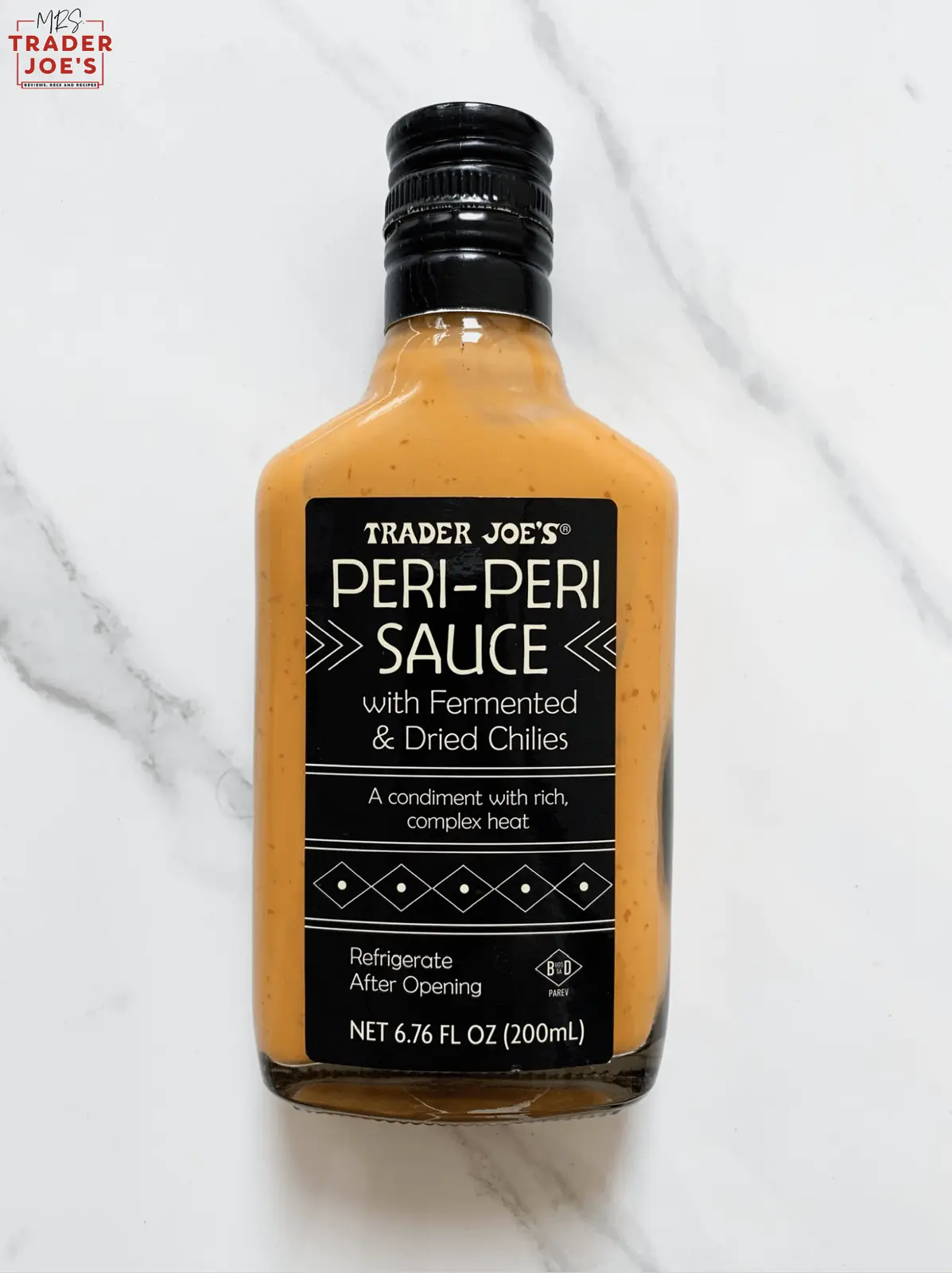 A bottle of Trader Joe's Peri-Peri Sauce with fermented and dried chilies sits on a white marble surface. The label…