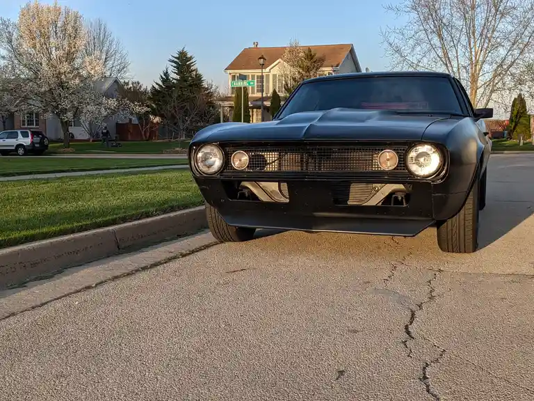 A matte black classic American muscle car, likely a Ford Mustang, is parked on a residential street with its headlights on…