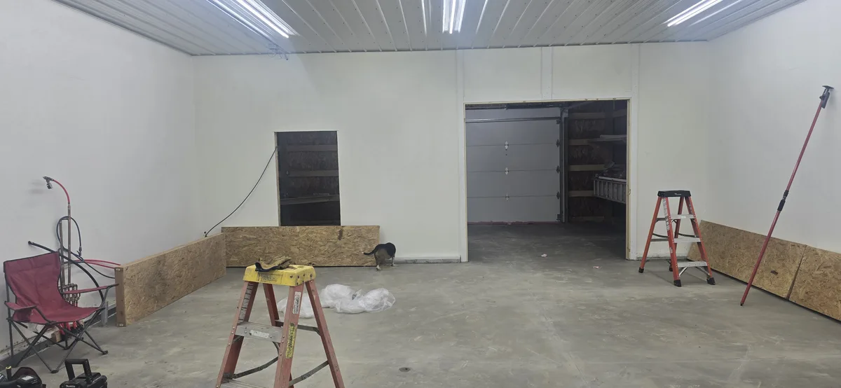 An empty, unfinished garage with white walls, a concrete floor, and construction tools like ladders and a red folding chair…