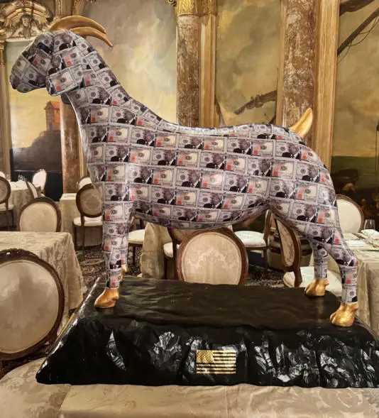 A large, ornate statue of a goat stands on a black pedestal in a luxurious dining room, its entire body covered in a…