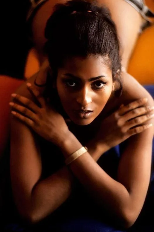 A woman with dark skin and her hair pulled back looks directly at the camera with a serious expression, her arms crossed…