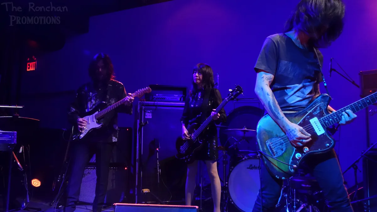 Three musicians are on stage under purple lighting, playing electric guitars and a bass guitar. The person on the right has…