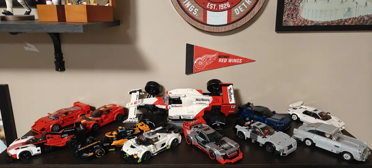A collection of various Lego car models, including race cars and sports cars, arranged on a dark wooden shelf. Behind them…