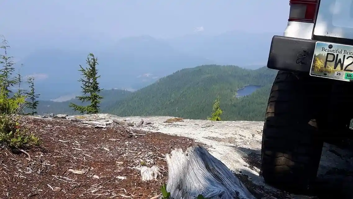 A vehicle's rear bumper and tire are visible on a rocky, forested mountain overlook, with a scenic view of distant forested…