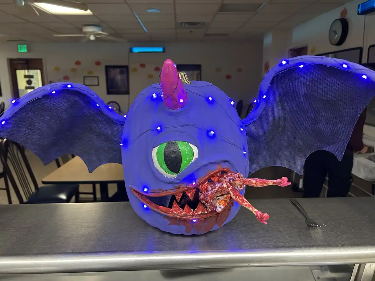 A large, blue-painted pumpkin is carved and decorated to resemble a dragon, featuring bat-like wings with blue LED lights, a…