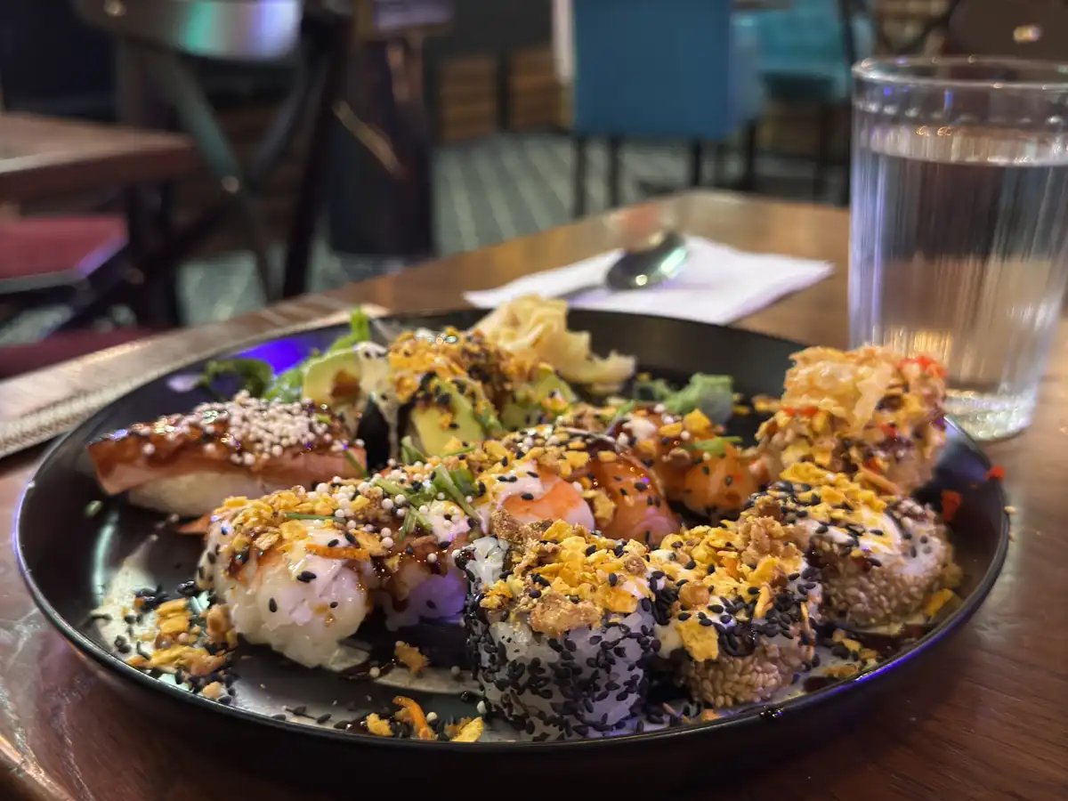 a close-up photo of a black plate piled high with various sushi rolls and nigiri, heavily garnished with black sesame seeds…