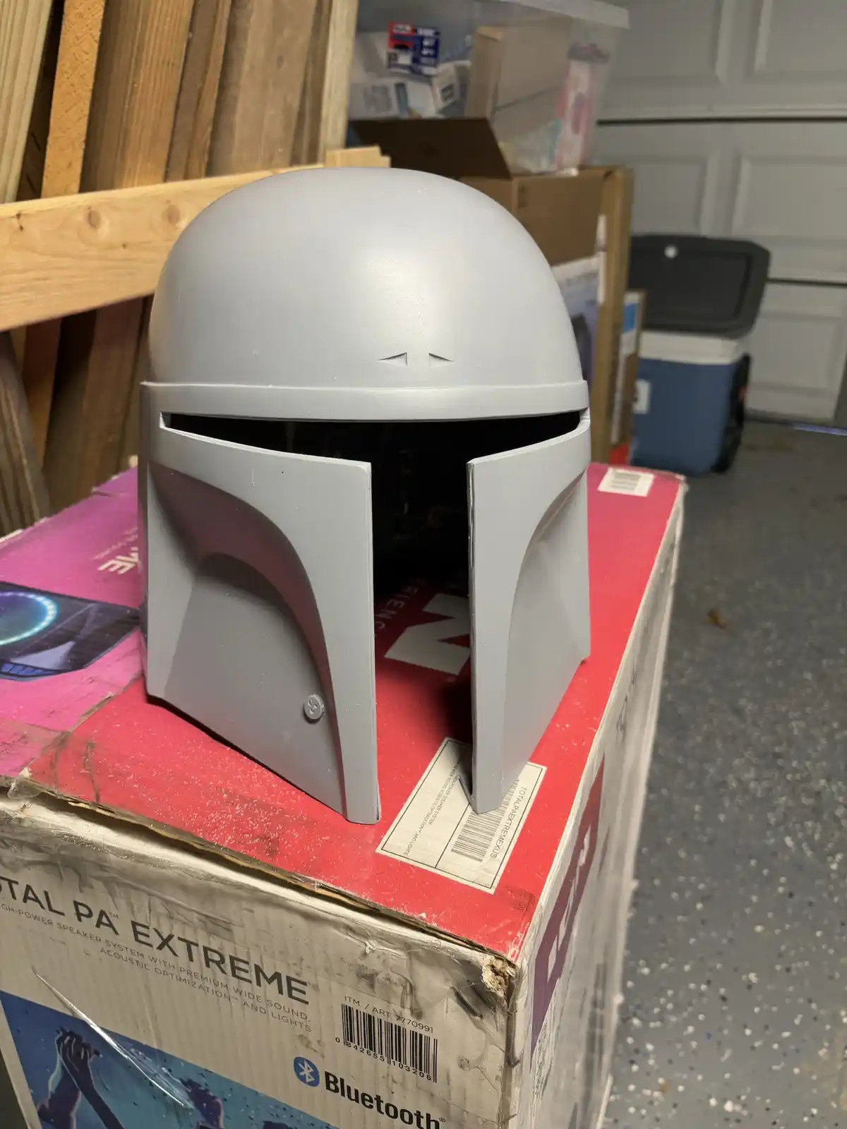 A gray-painted Mandalorian helmet, resembling The Mandalorian's iconic armor, sits atop a large red and white box labeled…