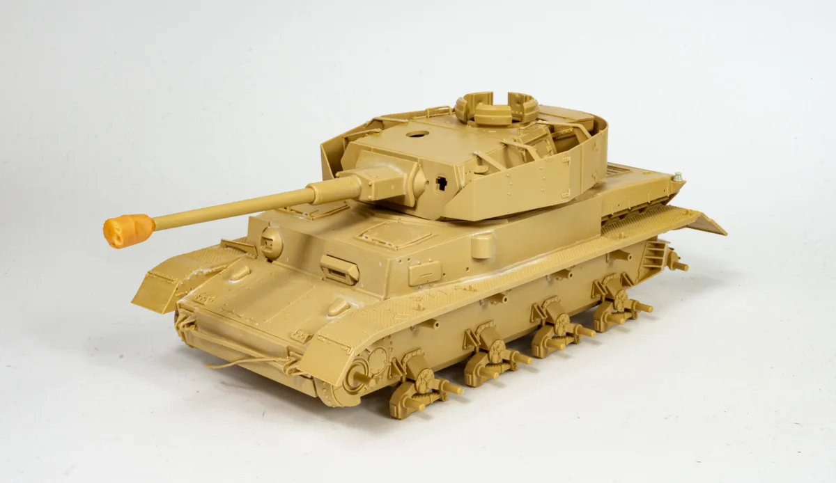 A tan plastic model tank kit, unassembled and unpainted, sits on a white surface. The tank has visible seams, missing parts…