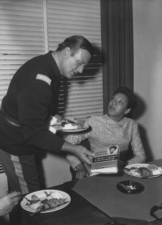 A black-and-white photo shows a man in a military-style uniform leaning over a table to serve food to a seated Black woman…