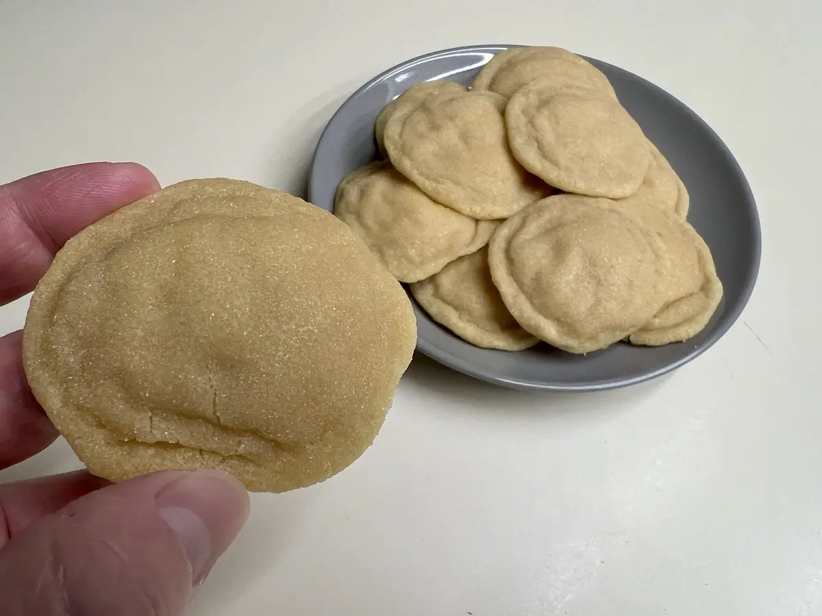 A hand holds a single round, sugar-dusted cookie in front of a gray bowl filled with many identical cookies. The cookies are…