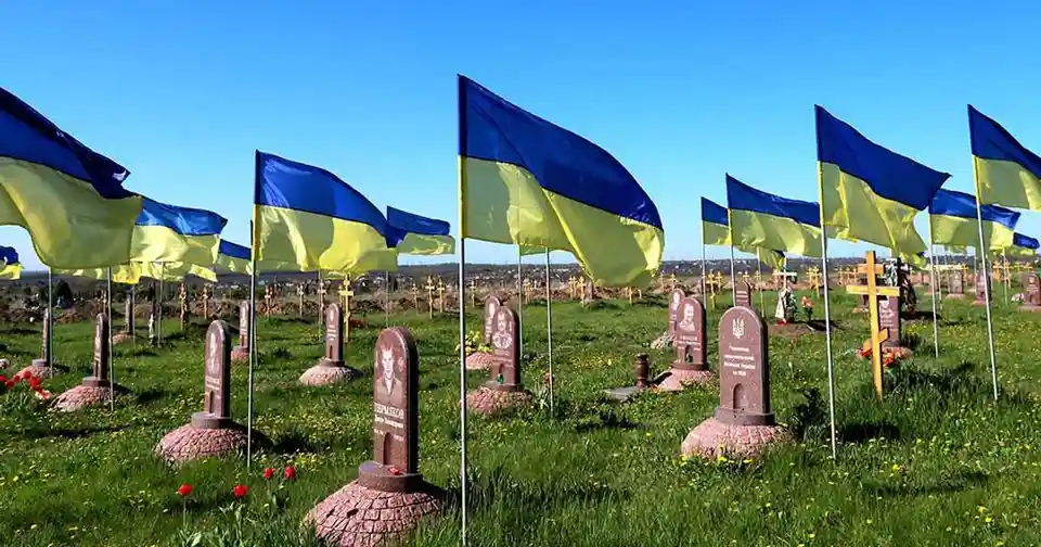 A wide shot of a cemetery under a clear blue sky, with numerous Ukrainian flags (blue and yellow horizontal stripes) waving…