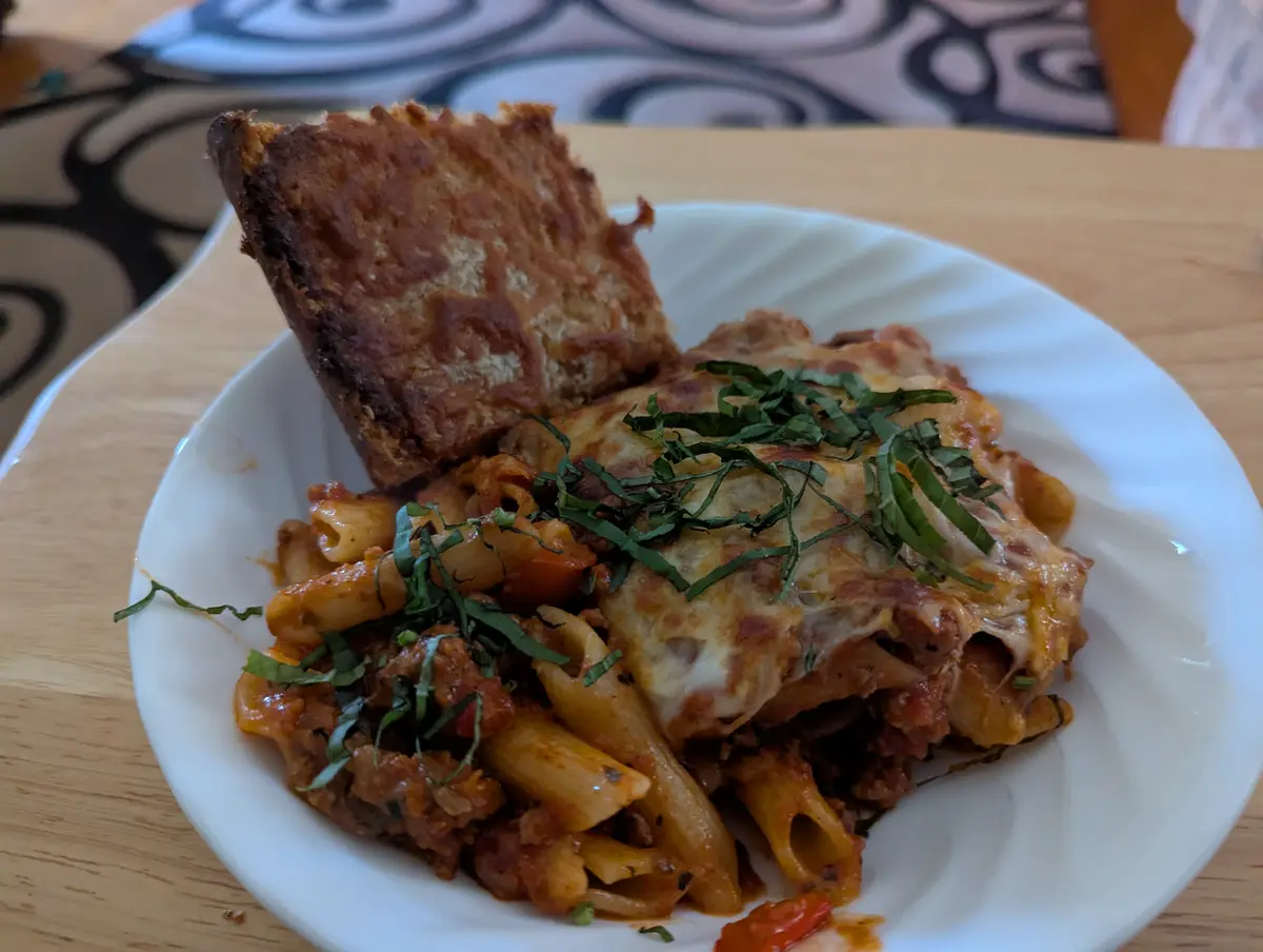 A white plate holds a serving of pasta with meat sauce and melted cheese, garnished with basil, alongside a piece of toasted…