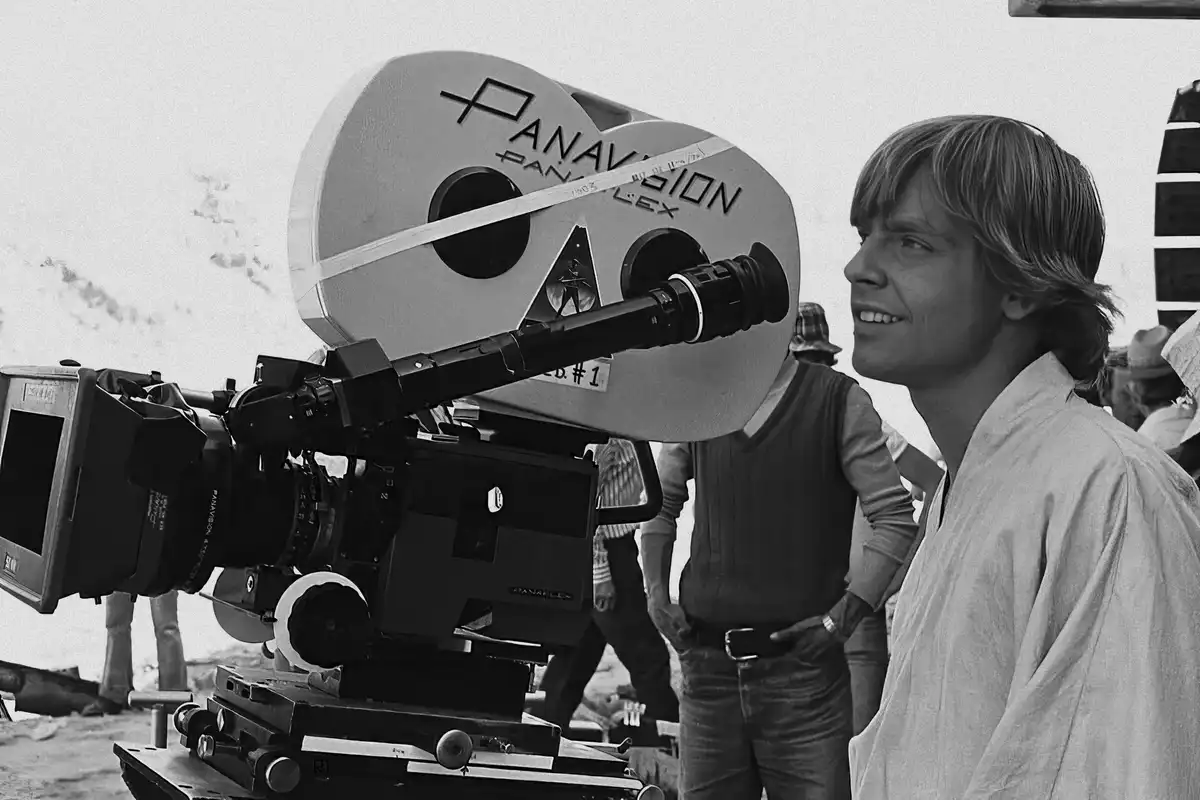 A black-and-white photo shows actor Mark Hamill on the set of Star Wars, smiling beside a large Panavision film camera. The…