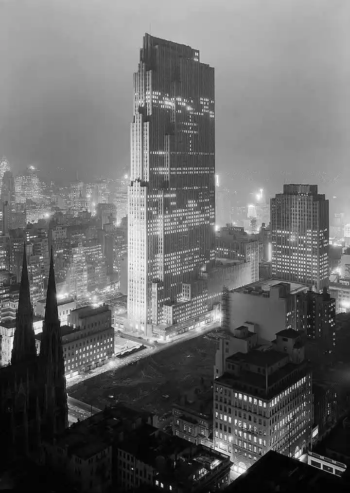 A black-and-white nighttime photograph of a tall skyscraper in a city, with its windows illuminated to form the shape of a…