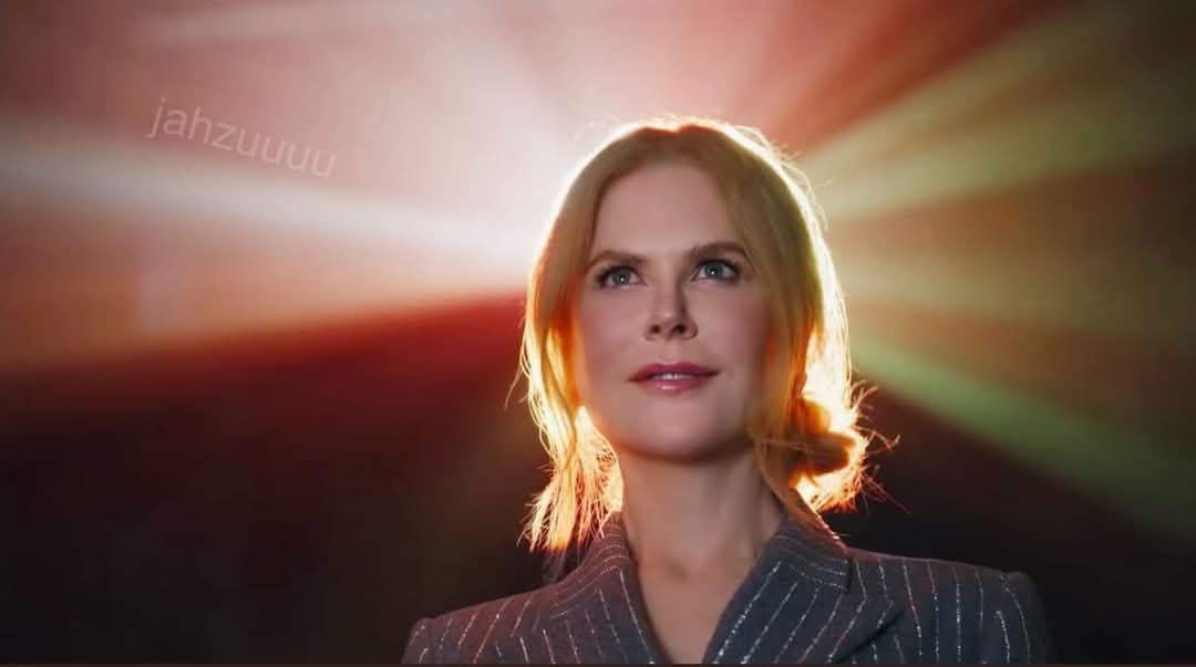 A woman with blonde hair and a pinstripe suit looks upward with a serene expression, illuminated by dramatic backlighting…