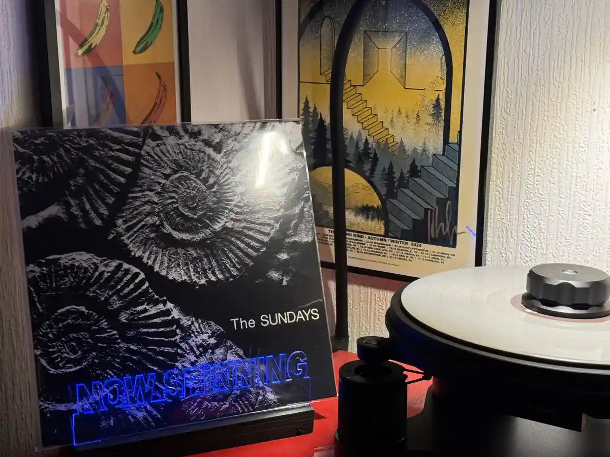A vinyl record album cover for 'The SUNDAYS' is displayed upright next to a turntable with a white record on it. The album…