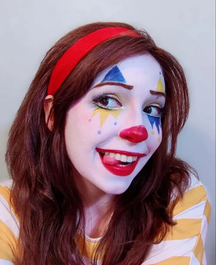 A woman with brown hair and a red headband wears full clown makeup including white face paint, blue and yellow triangles…