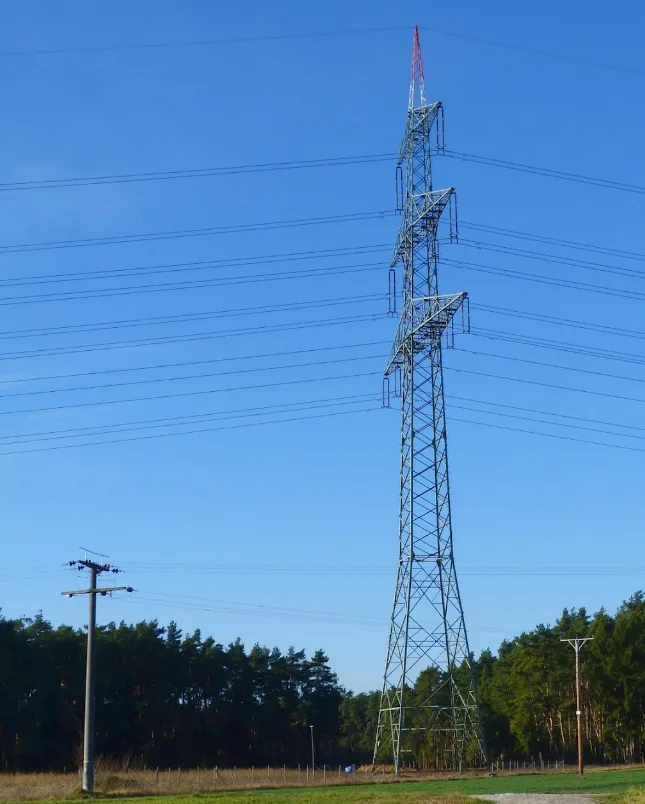 A tall, metal electricity pylon stands against a clear blue sky, with multiple power lines stretching across the frame. A…
