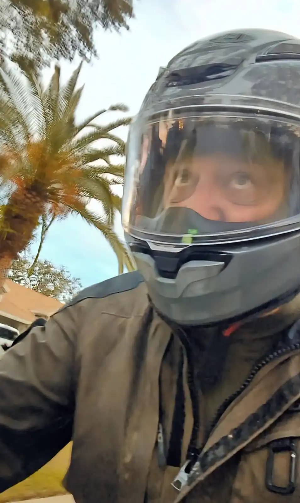 A person wearing a full-face motorcycle helmet and jacket looks directly at the camera with wide, startled eyes. The…