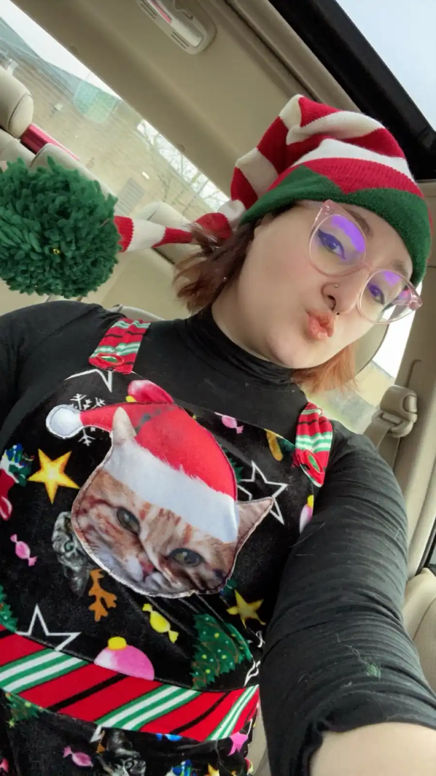 A person is taking a selfie inside a car, wearing a red-and-white striped elf hat and a black Christmas-themed shirt with a…