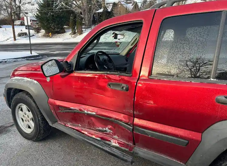 A red Jeep Liberty is parked on a snowy street, its side door and lower body heavily damaged with scraped paint and snow…