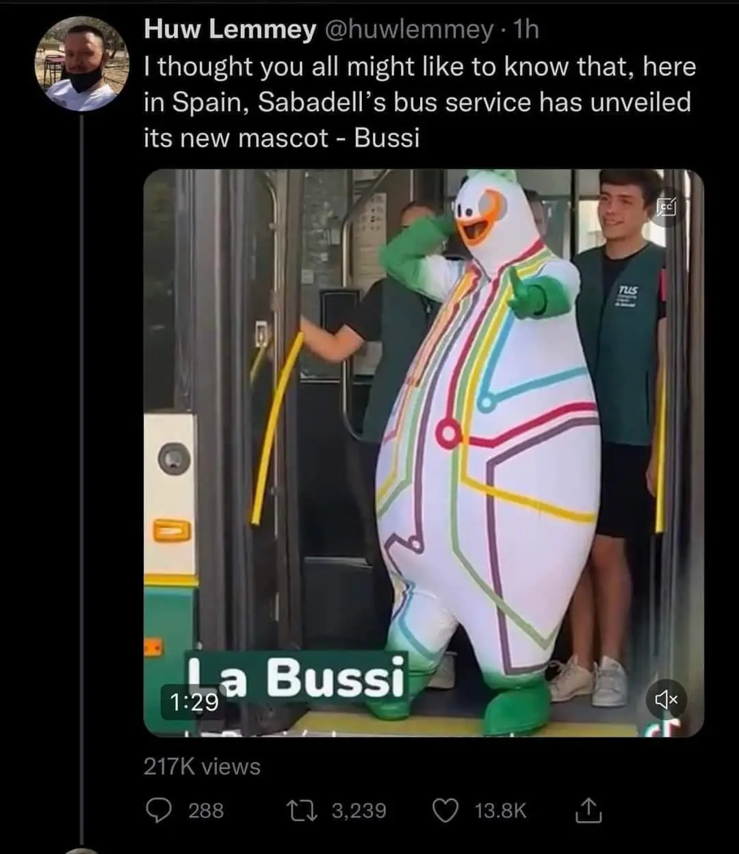 A person in a large, white, inflatable mascot costume with green feet, orange beak, and colorful circuit-like lines stands…
