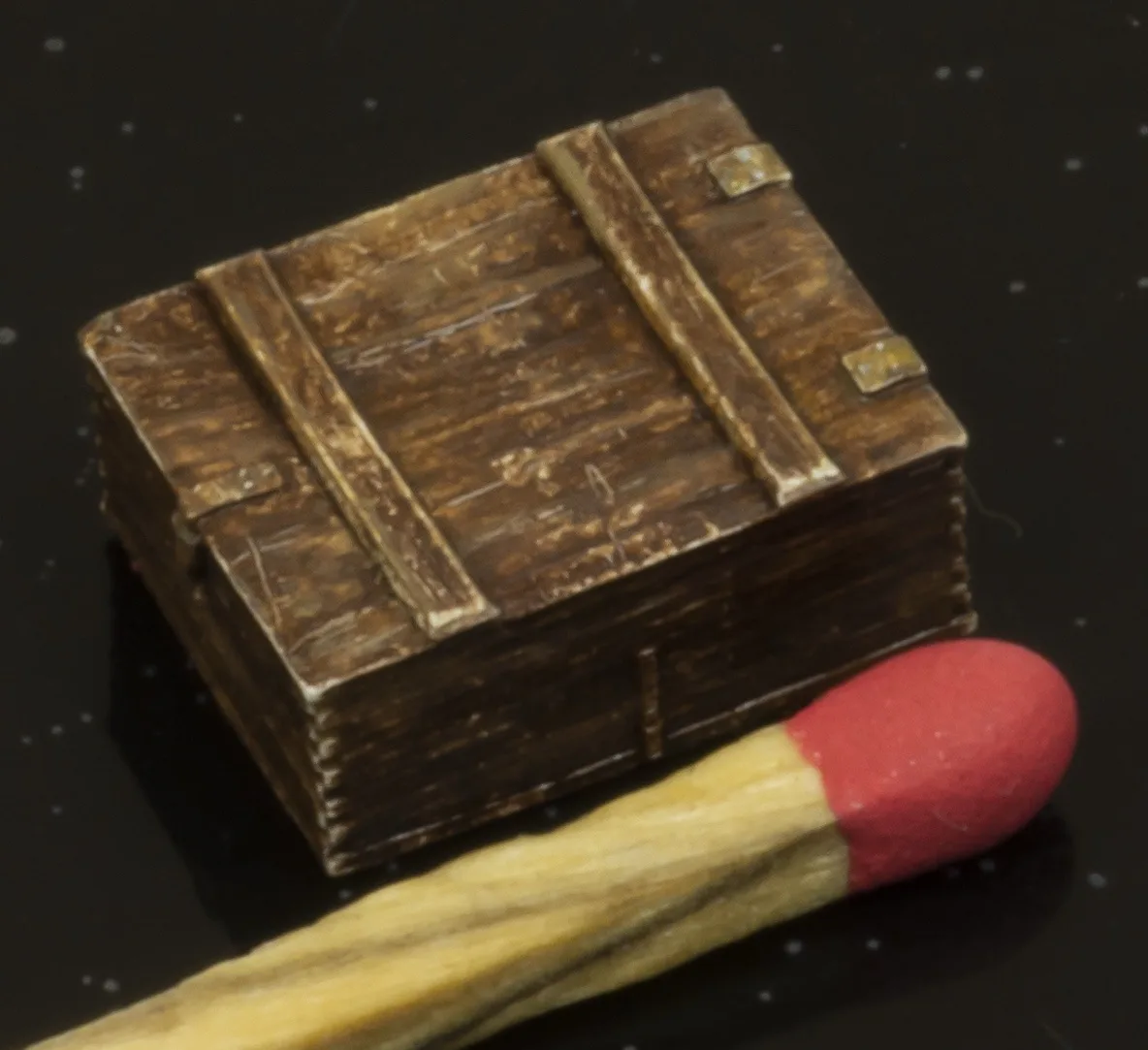 A small, weathered wooden treasure chest sits next to a single matchstick on a dark, speckled surface. The chest has metal…