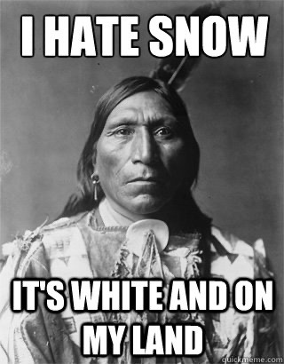 A black-and-white historical photograph of a Native American man with a stern, unamused expression, wearing traditional…
