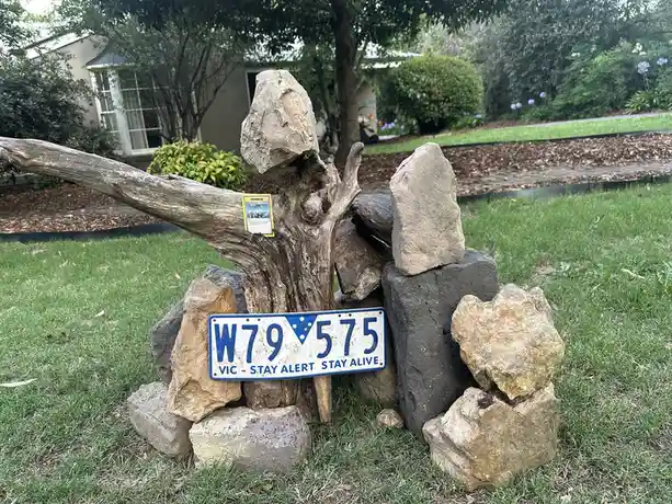 A Victorian license plate reading 'W79 575 VIC - STAY ALERT STAY ALIVE' is propped up among rocks and driftwood in a grassy…