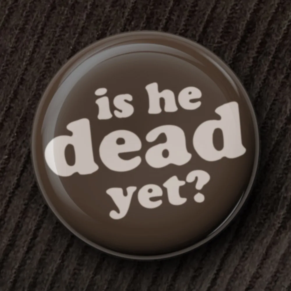 A close-up of a round, glossy brown button with white text that reads 'is he dead yet?' resting on a dark, textured fabric…