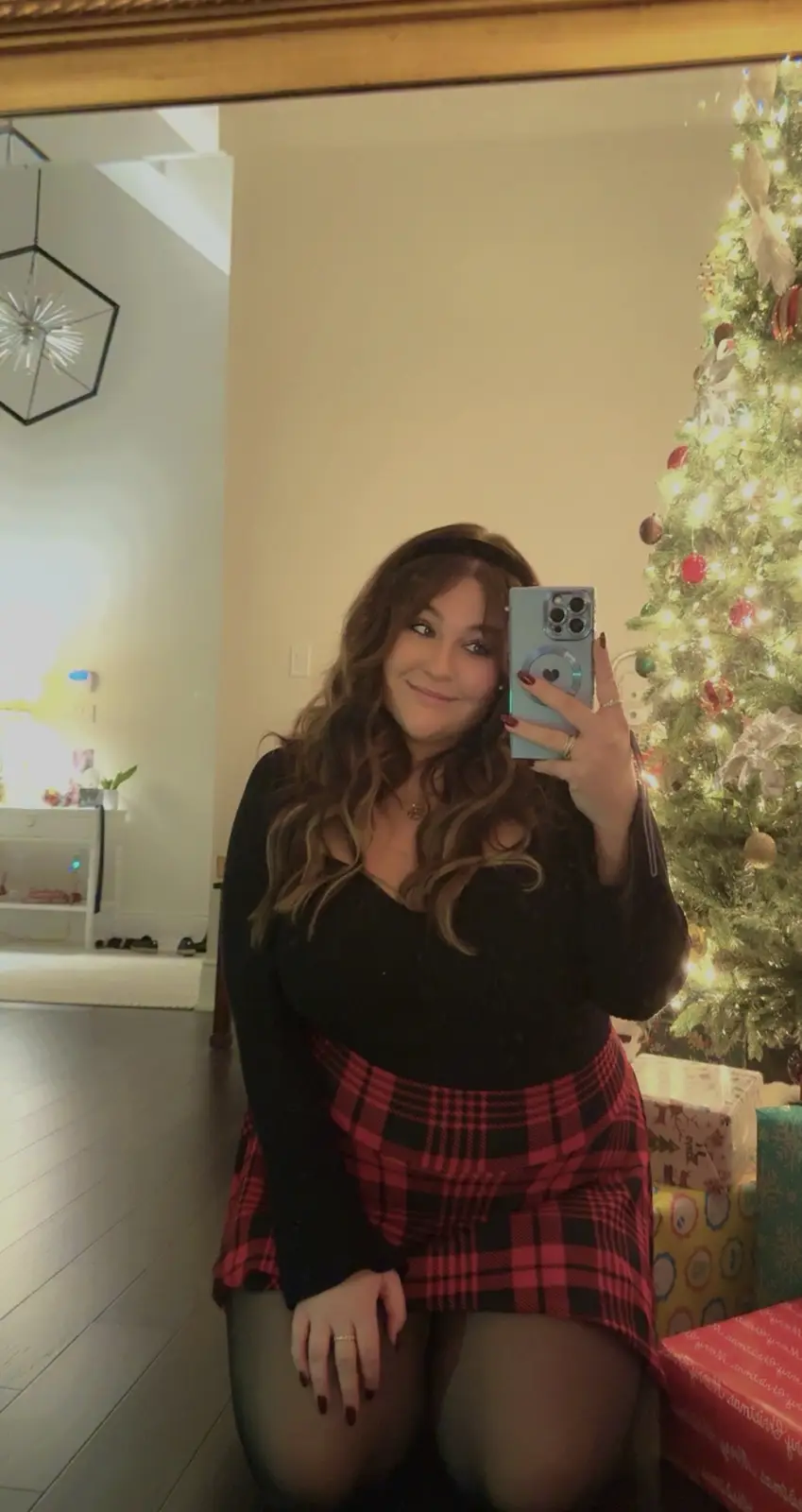 A woman takes a mirror selfie in a festive setting with a decorated Christmas tree behind her. She wears a black sweater and…