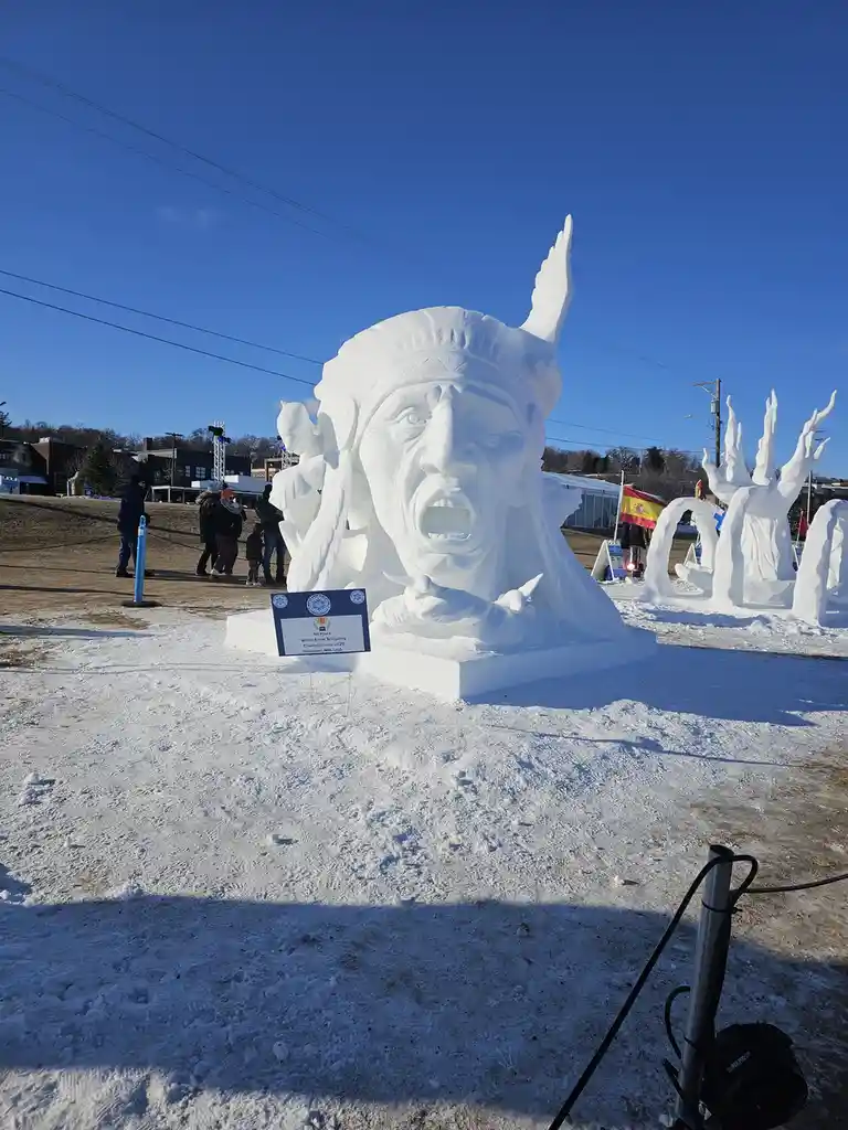 A large, detailed snow sculpture of a Native American chief with an open mouth and feathered headdress stands in a snowy…