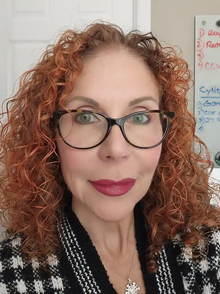 A woman with curly red hair and glasses looks directly at the camera with a slight smile, wearing a black and white…