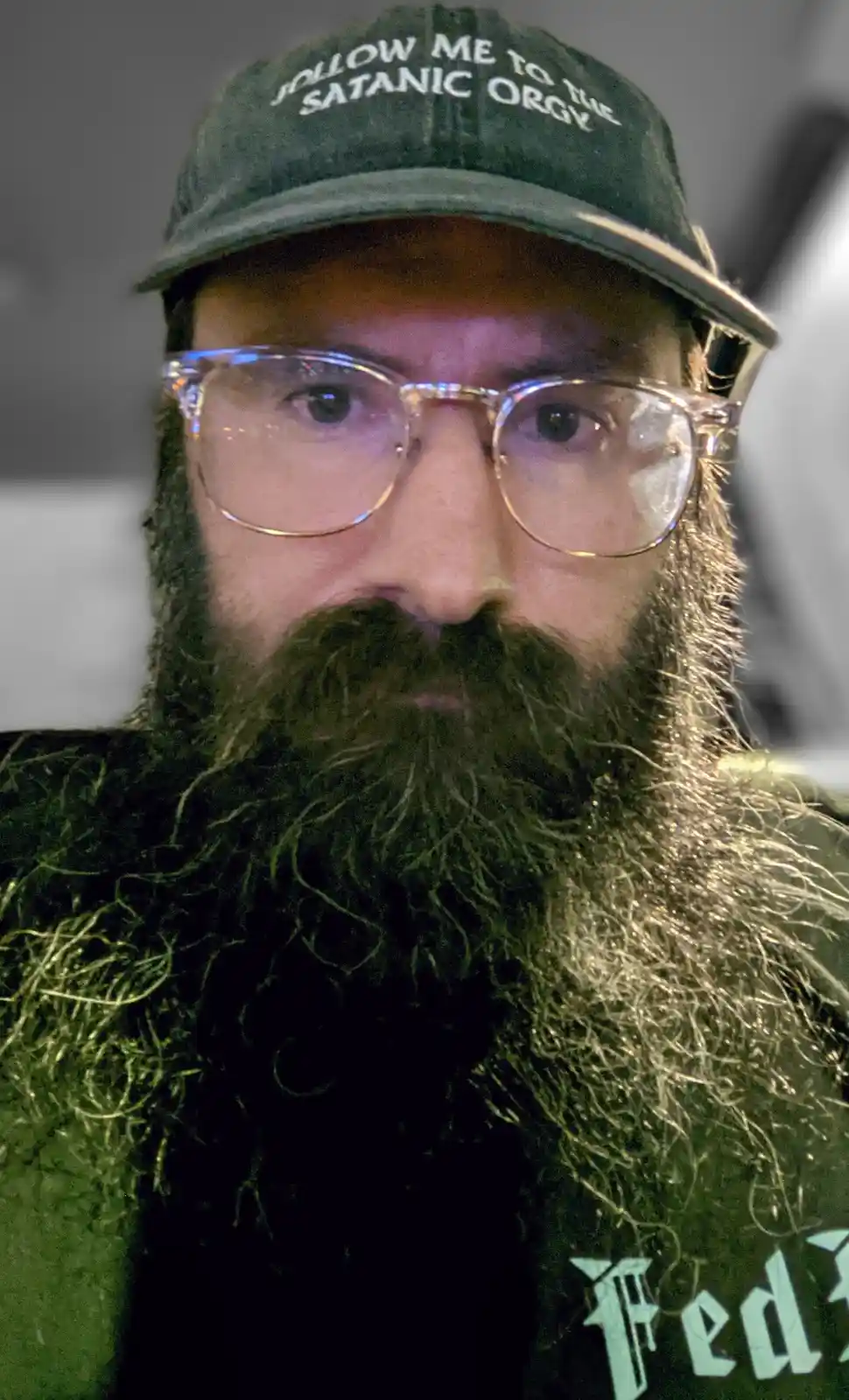 A close-up photo of a man with a long, dark beard and glasses, wearing a dark green baseball cap with white text that reads…