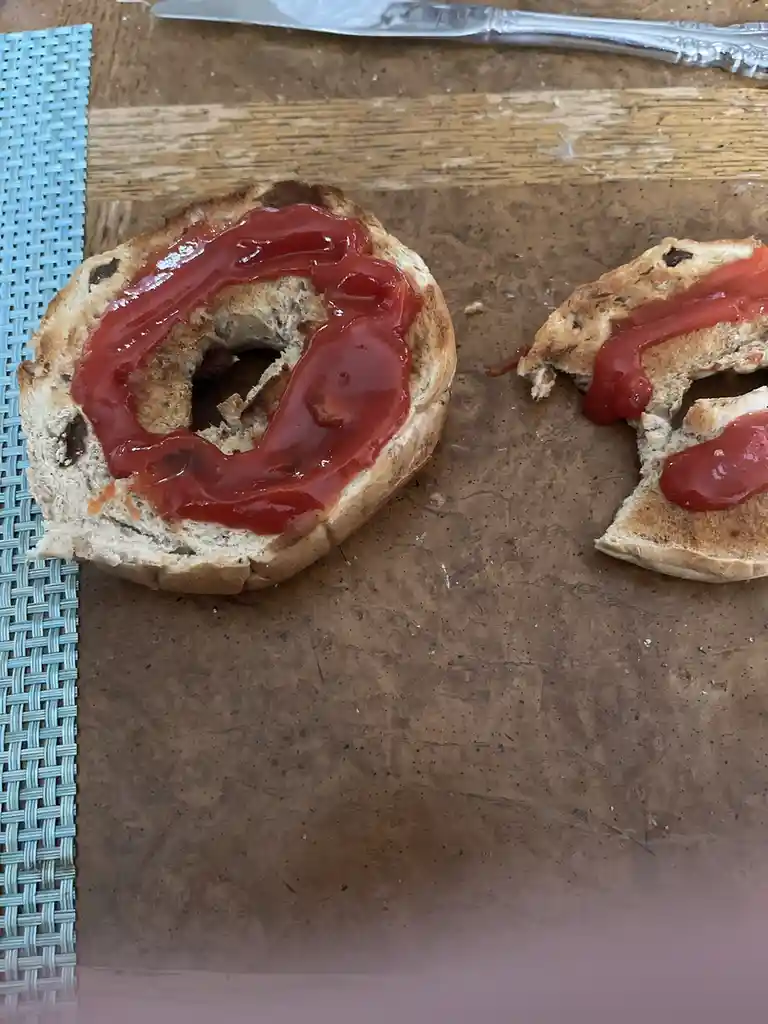 A split bagel with ketchup smeared on both halves sits on a wooden surface next to a blue woven placemat and a knife. The…