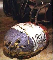A weathered, cracked, and painted helmet with red, white, and blue sections lies on the ground, with the number '1812'…