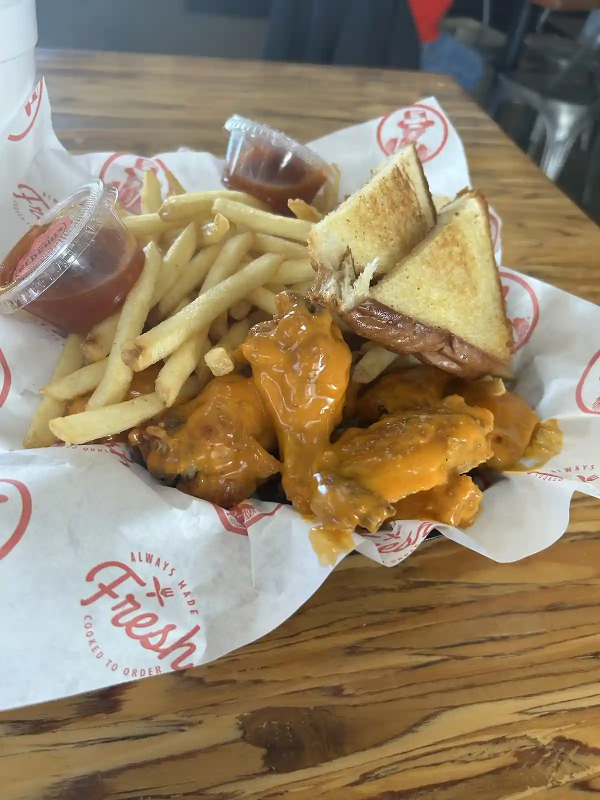 A close-up photo of a meal on a wooden table, featuring cheese-covered chicken wings, golden french fries, and a grilled…