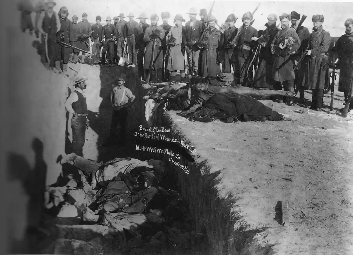 A historical black-and-white photograph shows a group of soldiers standing in the background holding rifles, while bodies of…