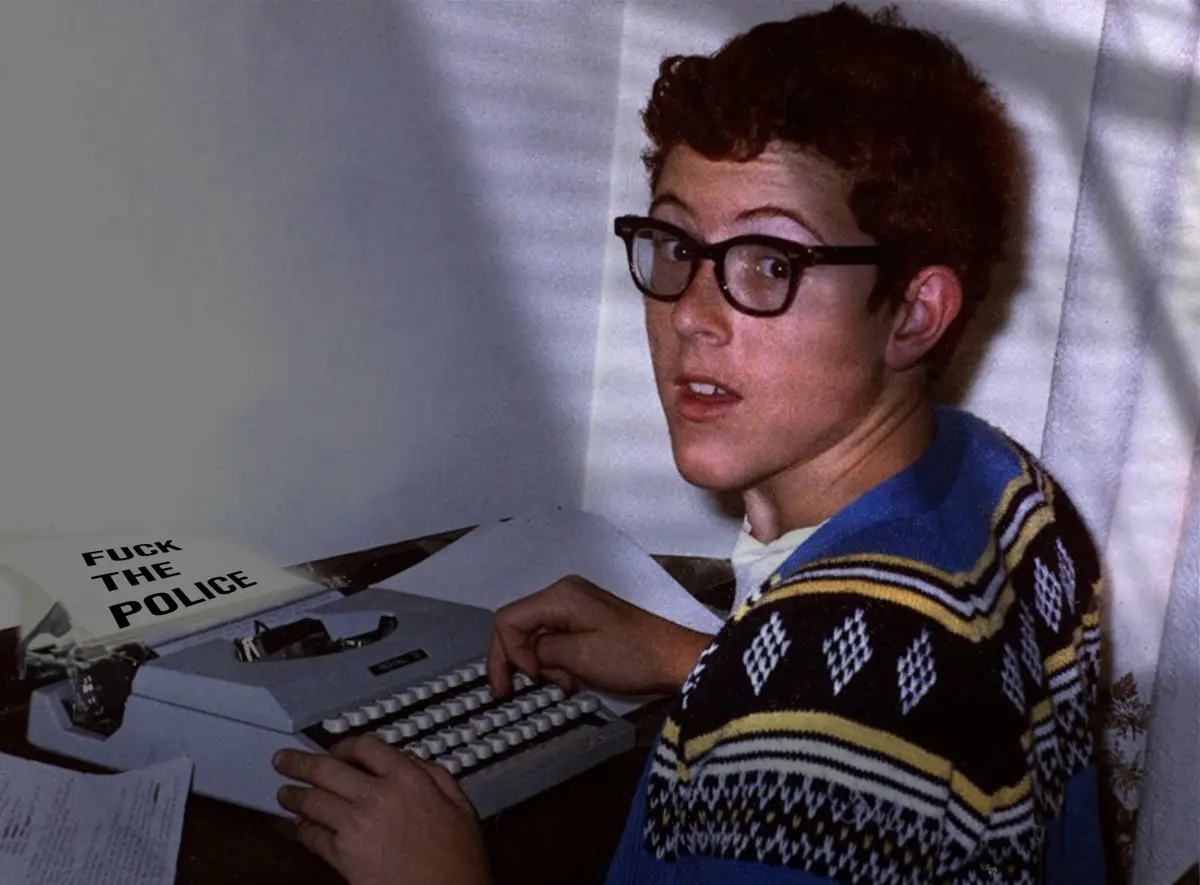 A young person with curly hair and glasses, wearing a patterned sweater, sits at a vintage typewriter and looks startled…