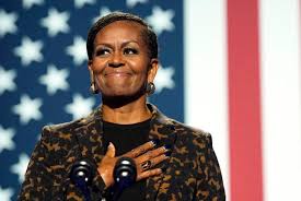 Michelle Obama stands with her right hand over her heart, smiling warmly, in front of a large American flag. She wears a…