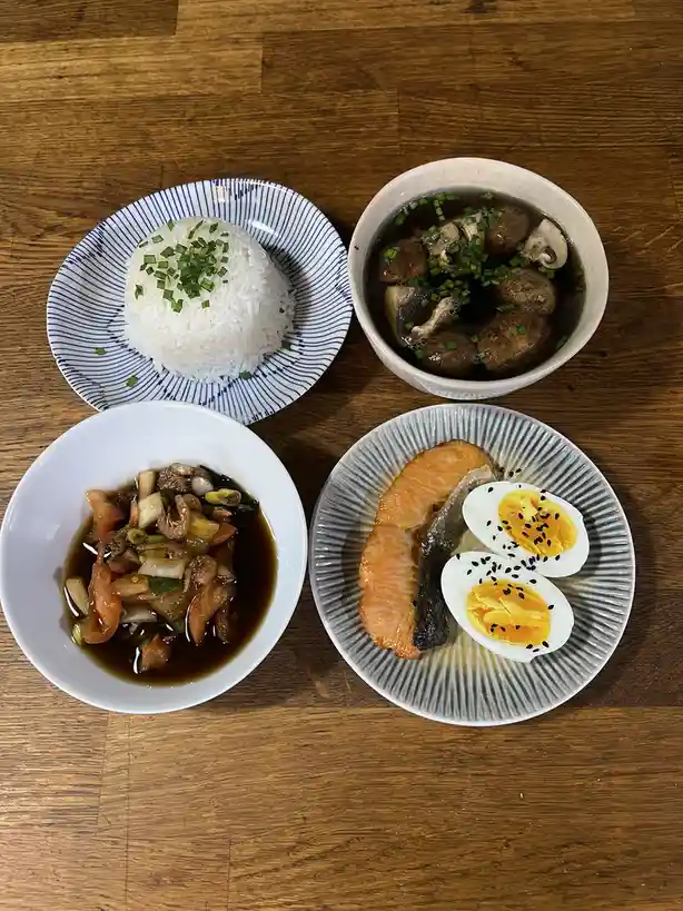 An overhead photo shows four dishes on a wooden table: a mound of white rice on a blue-striped plate, a bowl of mushroom…