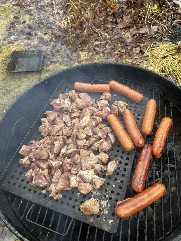 An overhead shot of a black charcoal grill outdoors, showing a pile of small, browned meat chunks on a perforated metal…